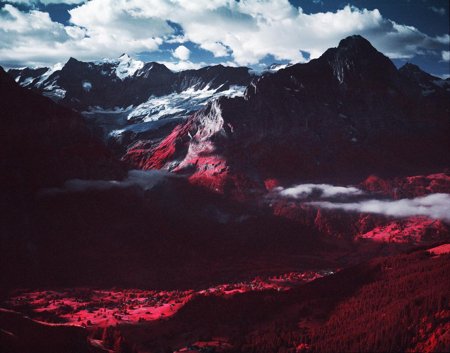 A mountain range with snow-capped peaks overlooks a valley with a small village and red foliage.