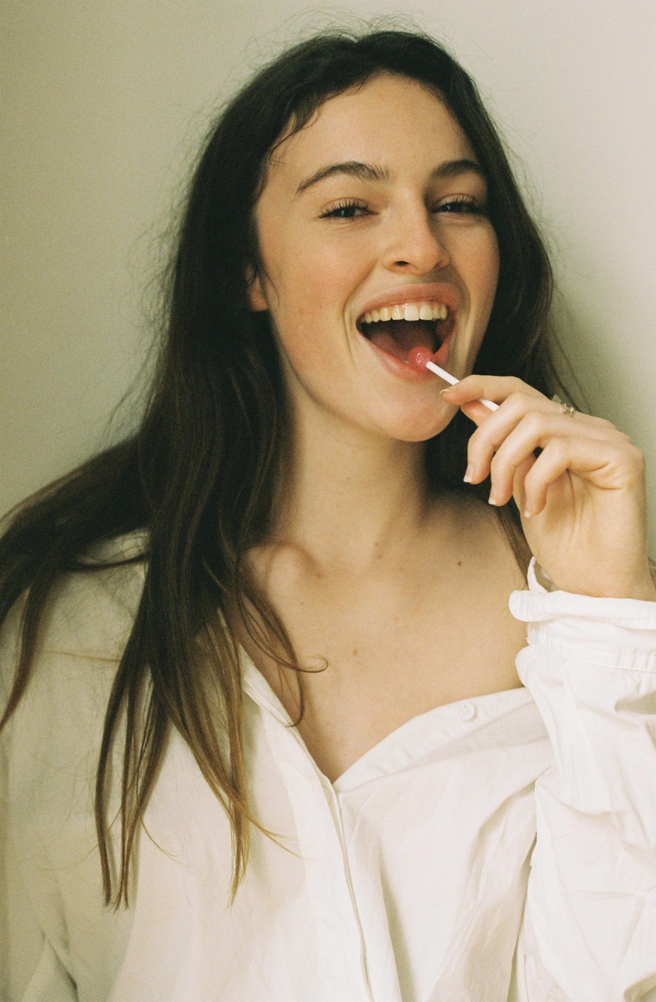 A woman with long hair and a white shirt laughs while holding a lollipop in her mouth.