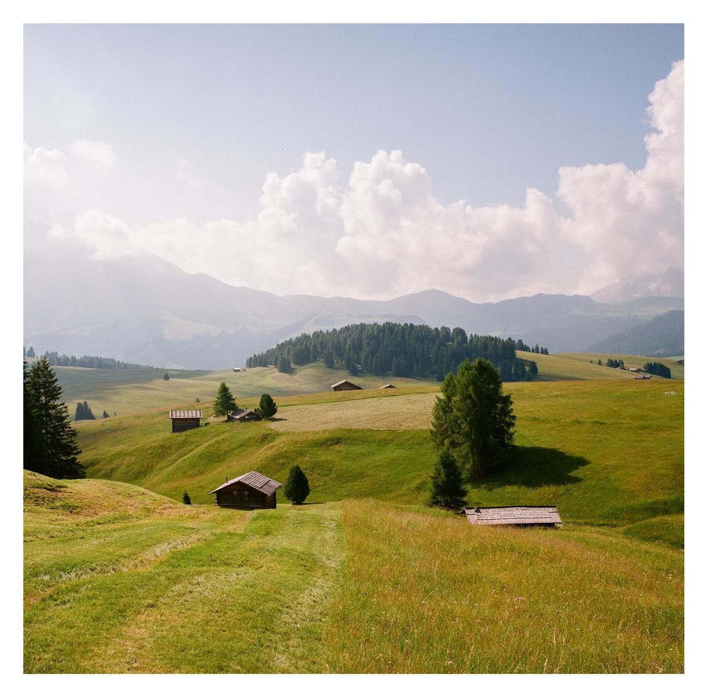 Small wooden huts are scattered across a green, hilly landscape with mountains and a forest in the background under a cloudy sky.