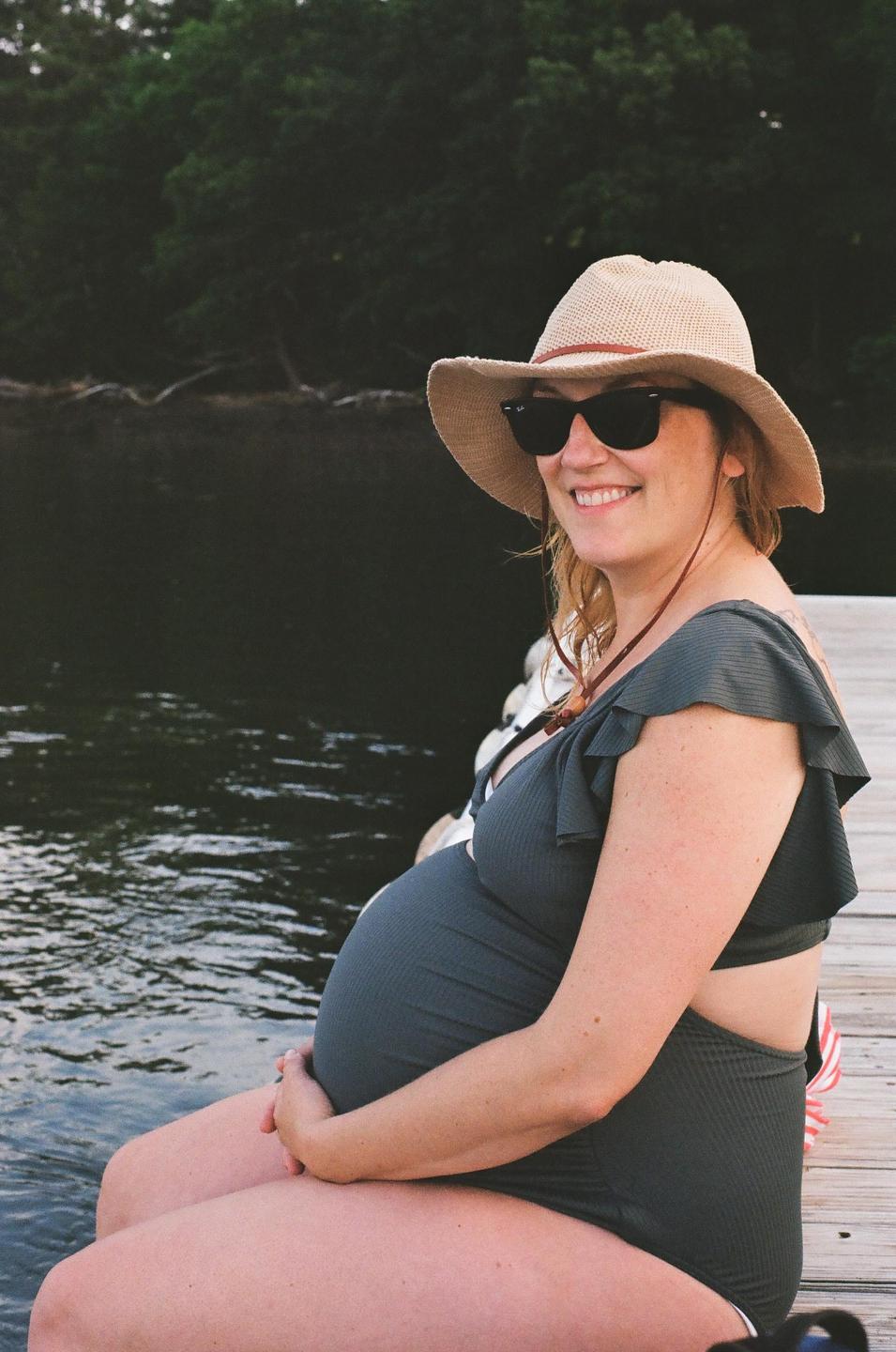 A pregnant woman wearing a sun hat and sunglasses sits on a wooden dock next to water with trees in the background.