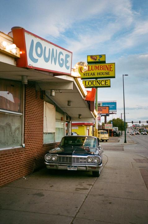 A vintage car is parked on a sidewalk next to a brick building with a "Lounge" sign.