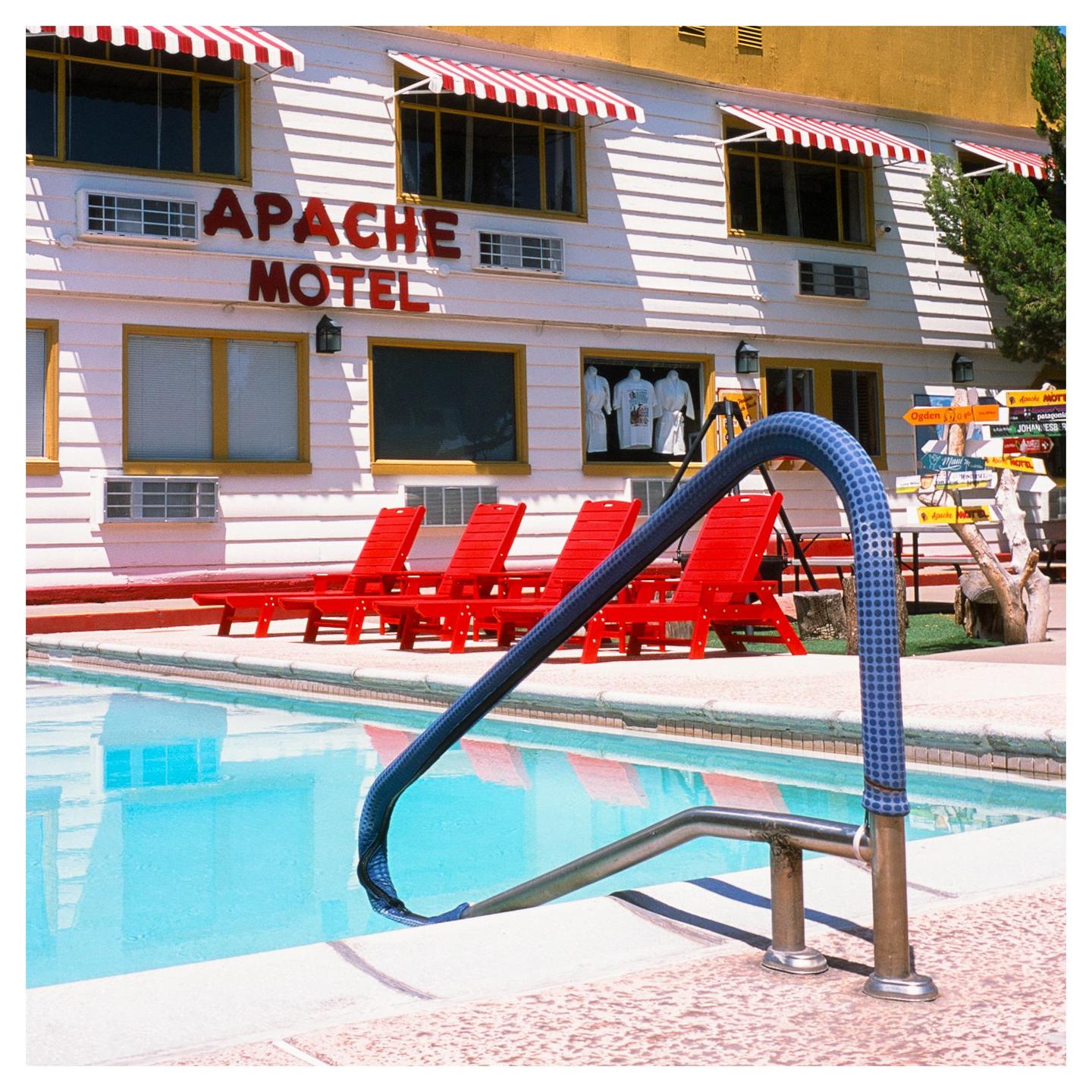 A swimming pool with a blue handrail is in front of the Apache Motel, which has red and white striped awnings and red lounge chairs.