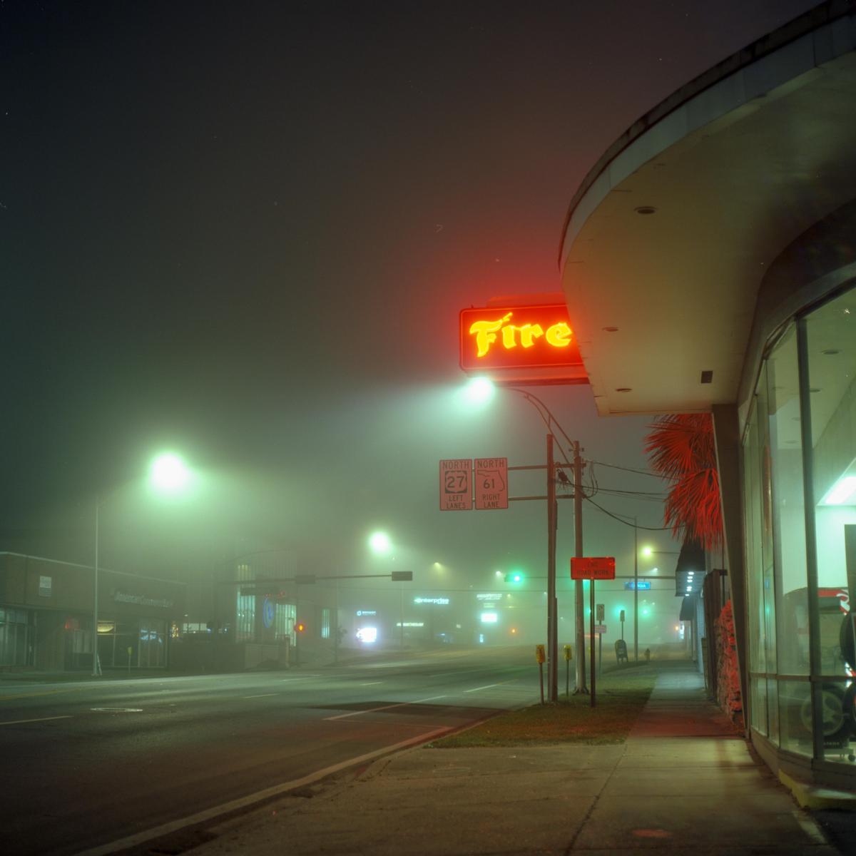 A neon sign reading "Fire" illuminates a foggy street at night, with a building and palm tree visible on the right.