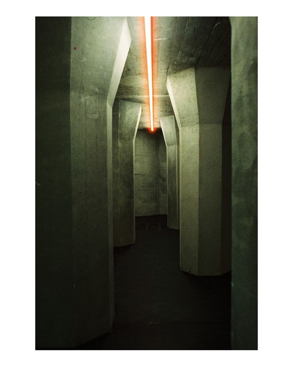 A long, narrow concrete corridor is illuminated by a single orange neon light running along the ceiling.