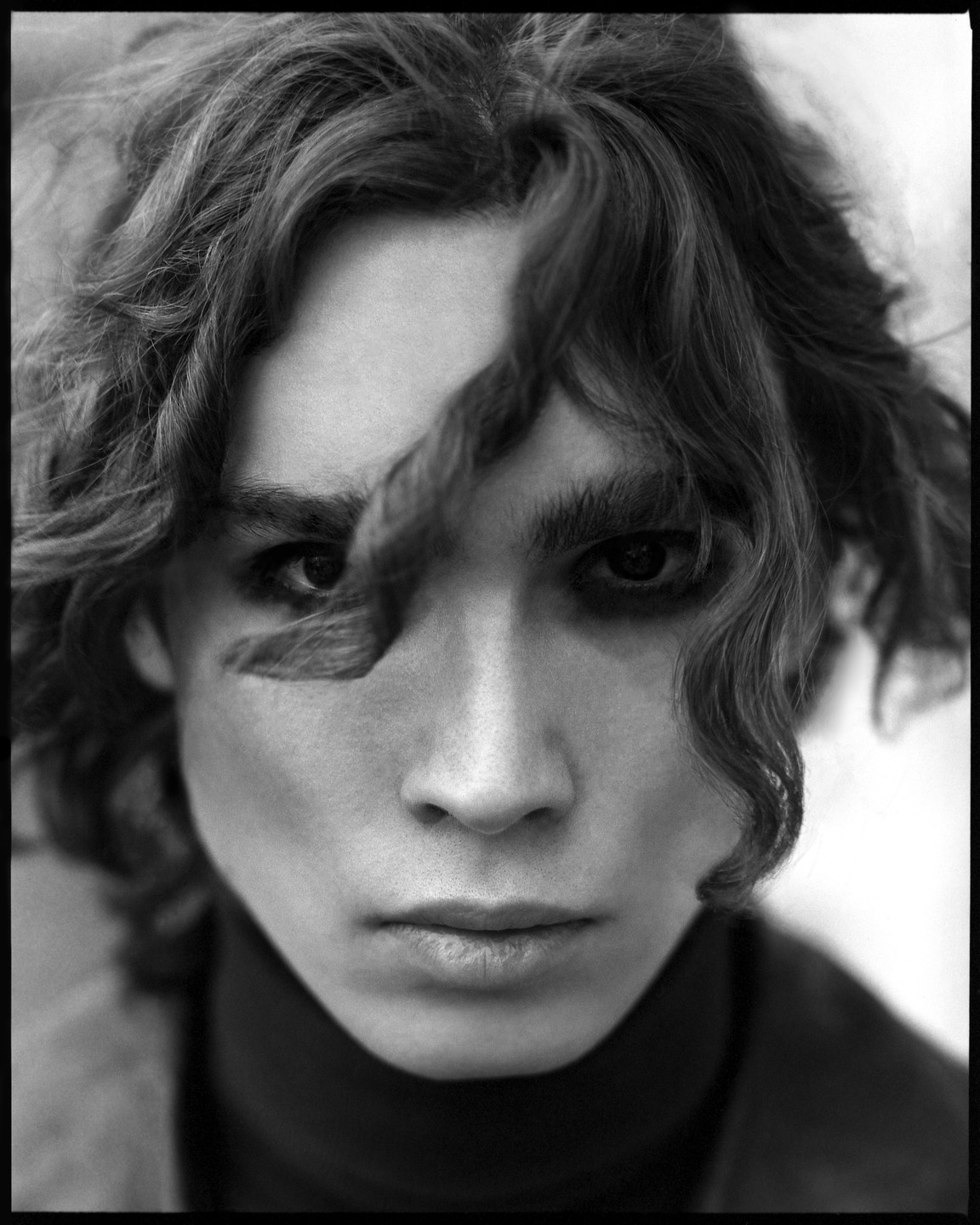 A person with curly hair and dark eye makeup looks directly at the viewer.