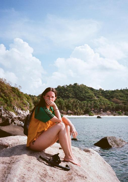 A woman sits on a large rock by the ocean with a tropical island in the background.