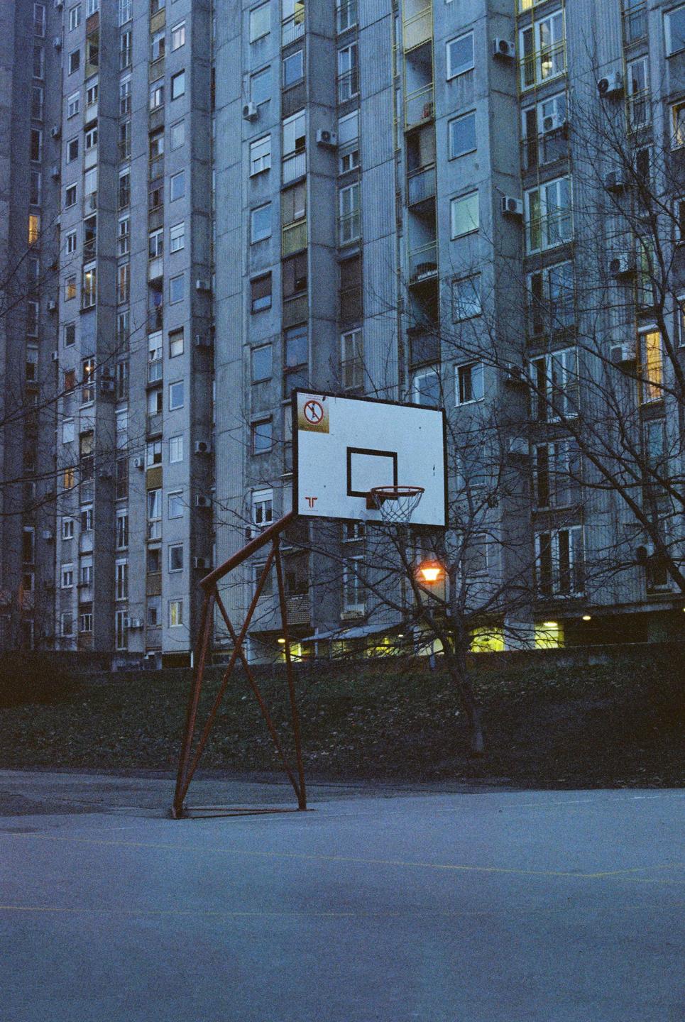 A basketball hoop stands on a court in front of a tall apartment building at dusk.