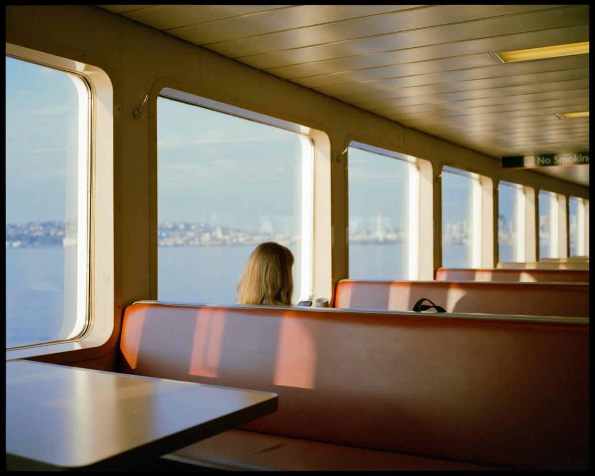 A woman sits on a ferry looking out at a city across the water during golden hour.