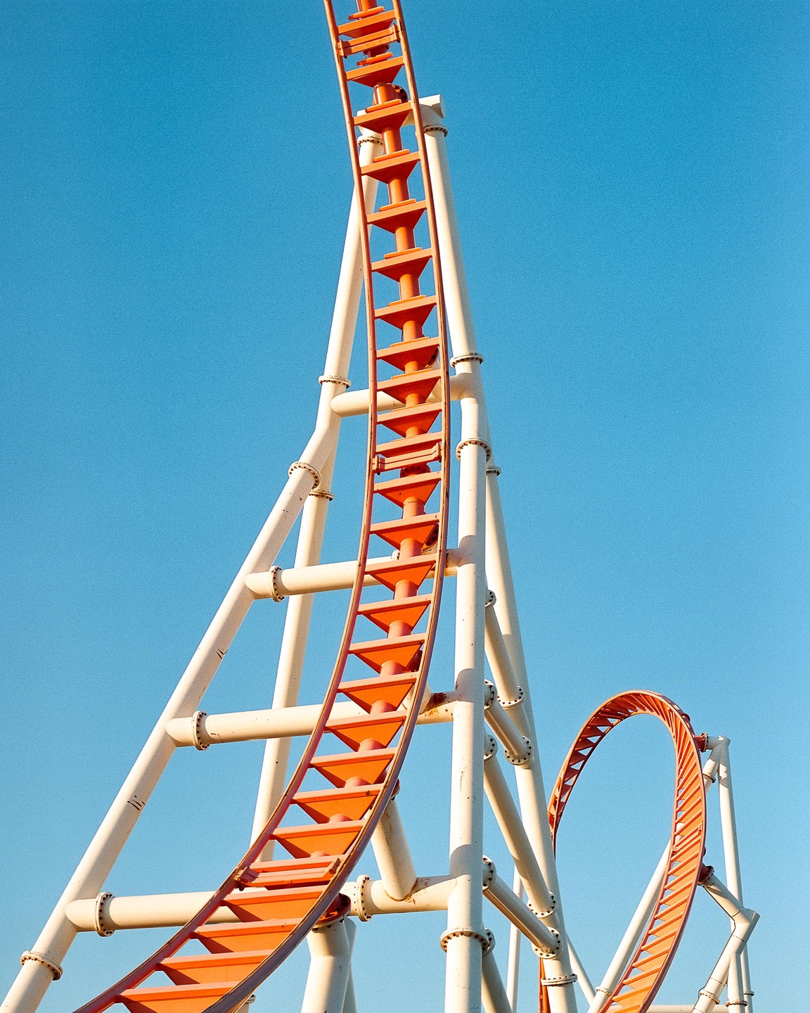 A bright orange roller coaster track with white supports curves against a clear blue sky.