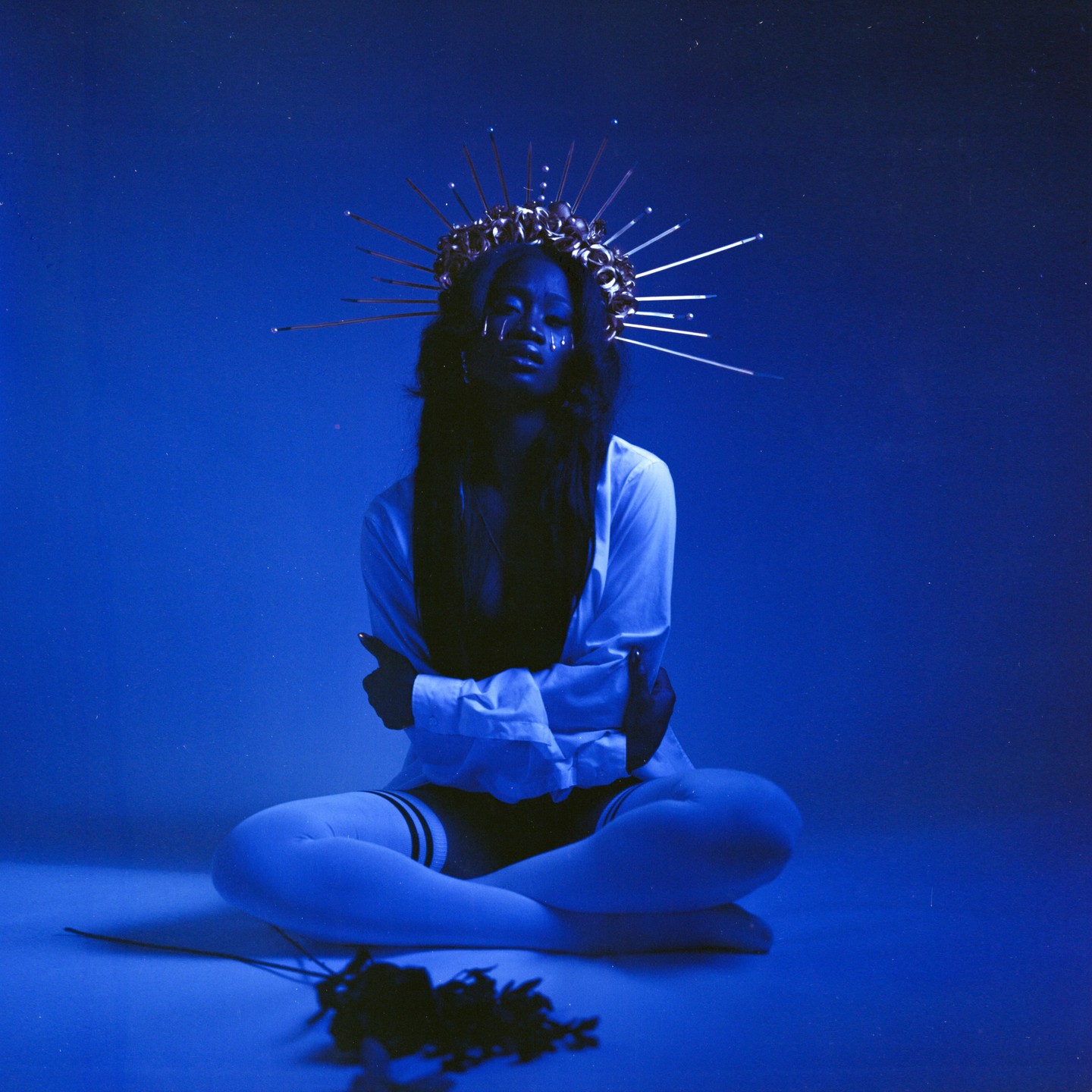 A woman wearing a crown and white shirt sits cross-legged on a blue surface with a bouquet of flowers in front of her.