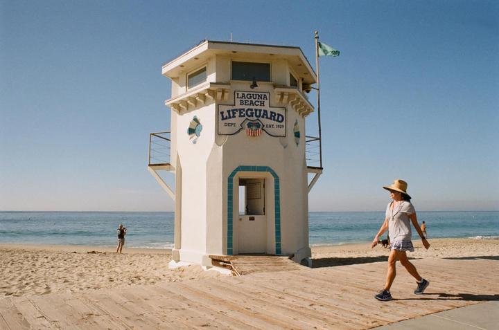 A woman walks past a white lifeguard tower on a wooden boardwalk next to the ocean.