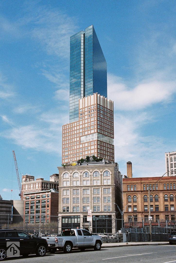 A tall, modern glass skyscraper rises above older brick buildings and parked cars on a city street.