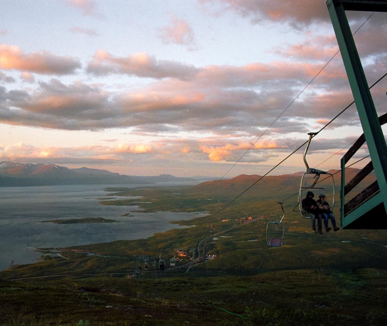 A couple rides a ski lift over a vast landscape with a lake and mountains under a cloudy sunset sky.