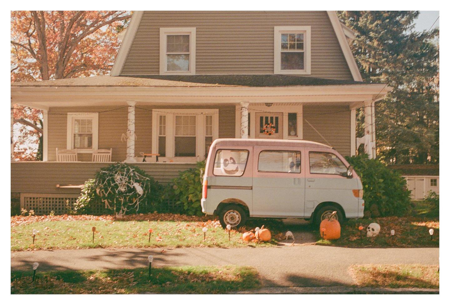 A two-tone van is parked in front of a house decorated for Halloween with pumpkins and skeletons.