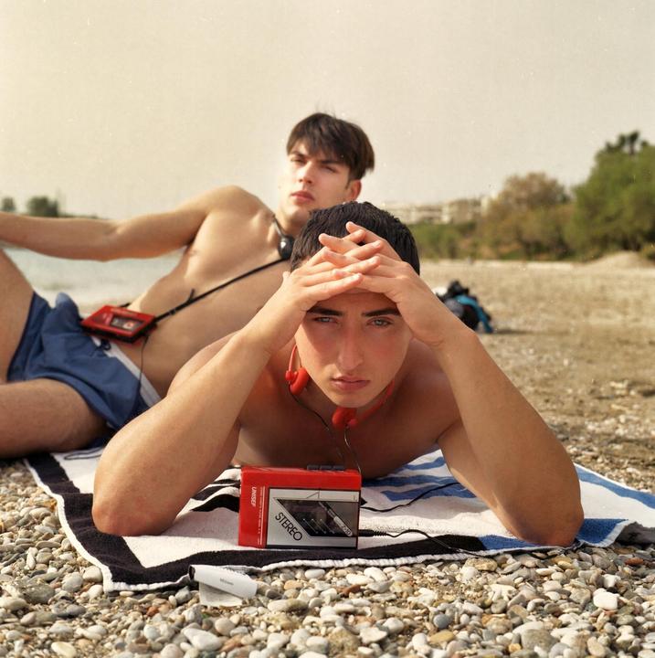 Two boys are lying on a rocky beach with a red stereo cassette player and headphones.