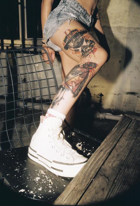 A person wearing denim shorts and white high-top sneakers stands with one foot on a chair, revealing tattooed legs.