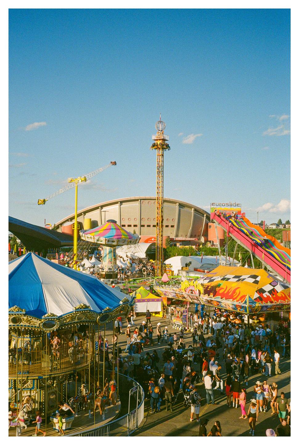 A large crowd of people gathers around various rides at a fairground under a clear blue sky.