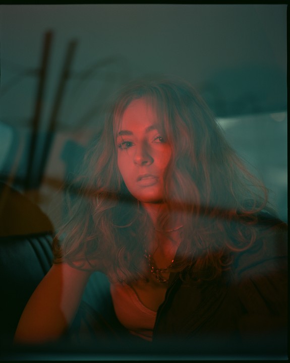 A woman with long wavy hair looks directly at the viewer from inside a vehicle, illuminated by red and green light.