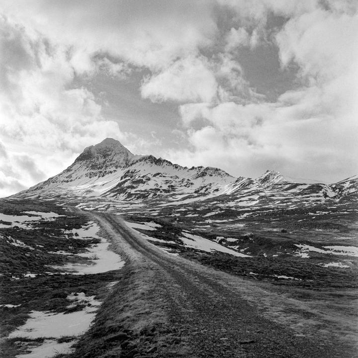 A dirt road leads towards a snow-capped mountain under a cloudy sky.