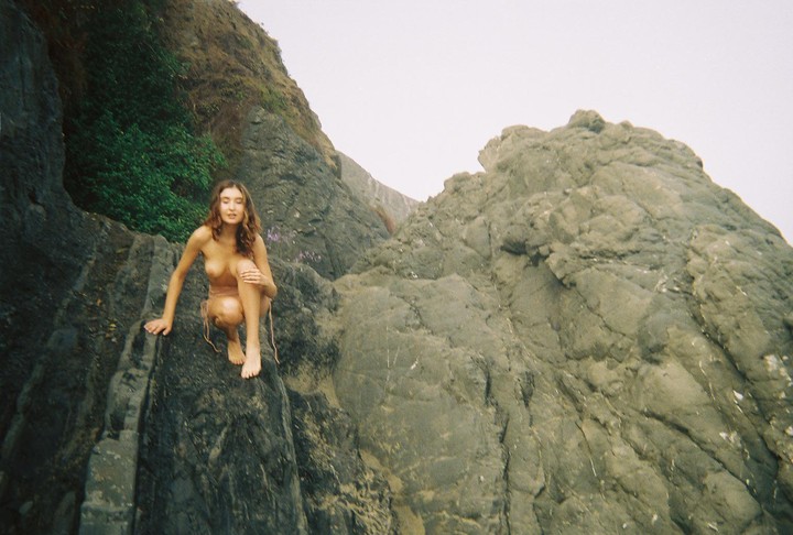 A woman crouches on a rock face with green foliage to her left and a large rock formation to her right.