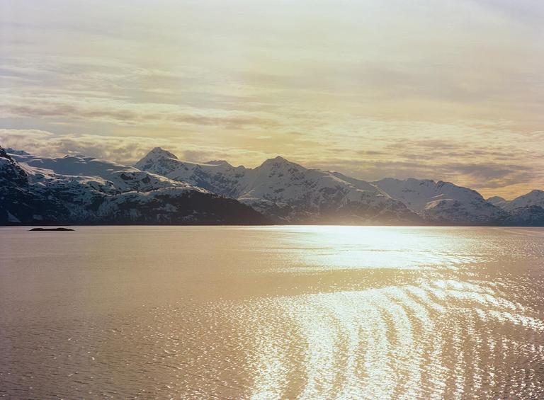 Sunlight reflects on the water in front of snow-capped mountains under a cloudy sky.
