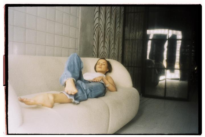 A woman in jeans and a white top is lying on a white sofa with her eyes closed.