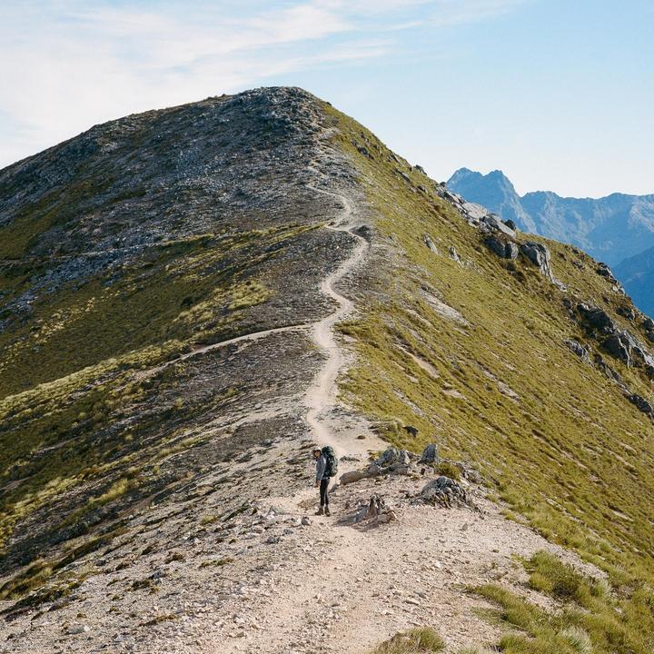 A person with a backpack stands on a winding trail along a mountain ridge under a clear sky.