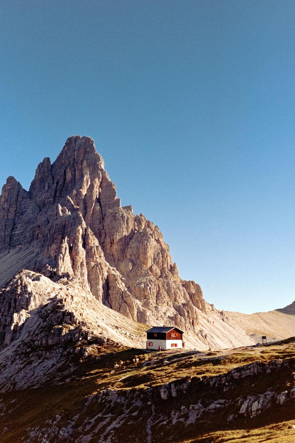 A small hut with a red roof sits at the base of a large, jagged mountain under a clear sky.