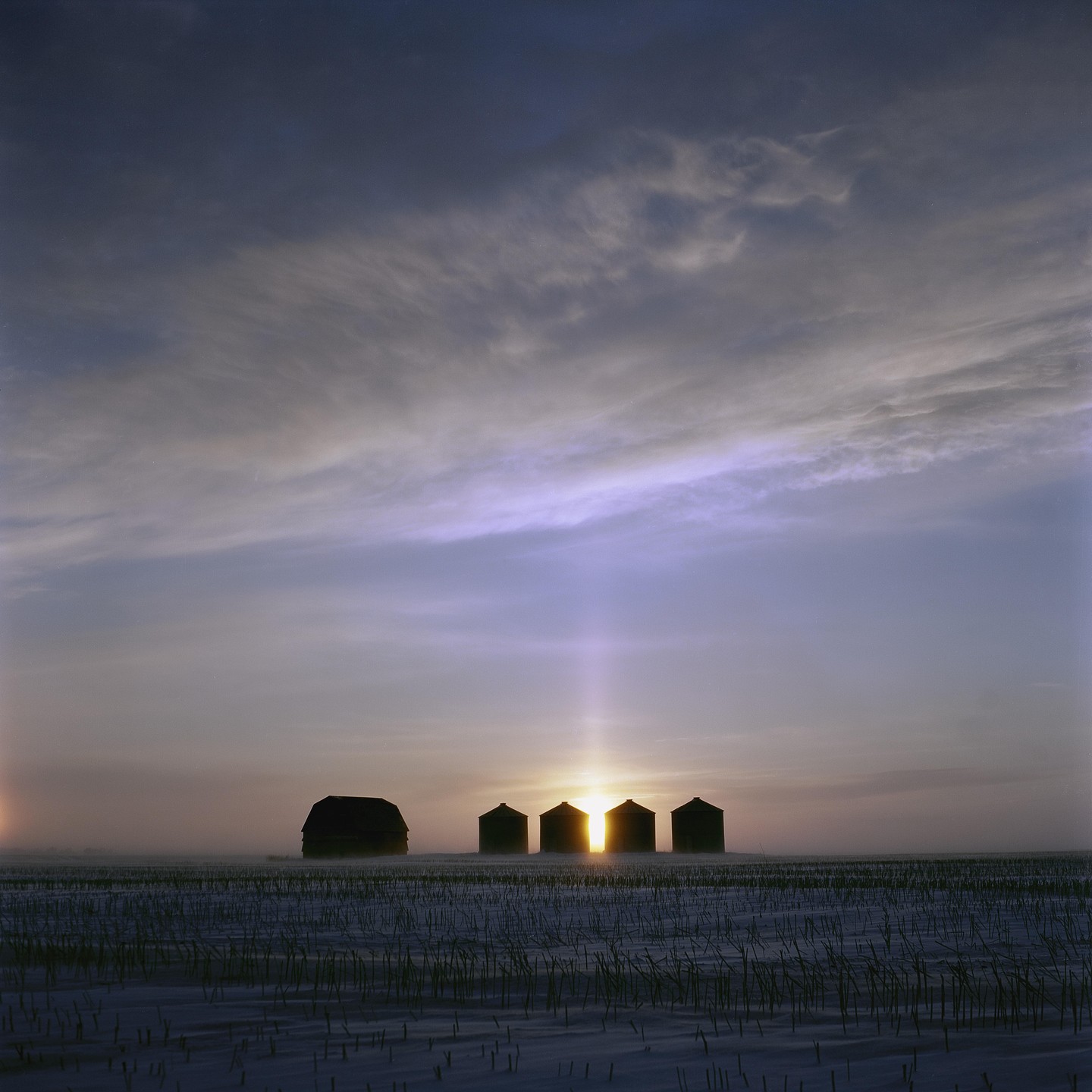 The sun rises behind a barn and four silos in a snowy field.