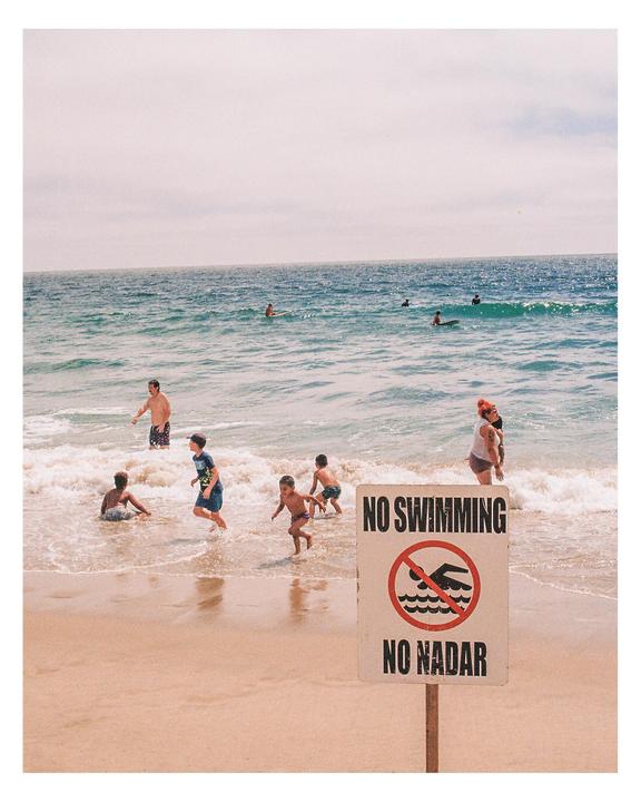 A "No Swimming" sign stands on a sandy beach with people in the ocean.