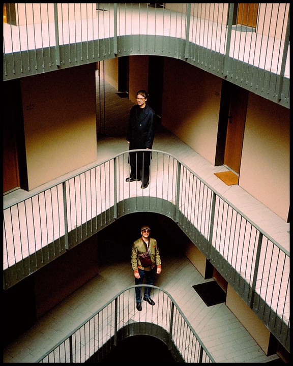 Two men stand on different levels of a building with curved hallways and railings.