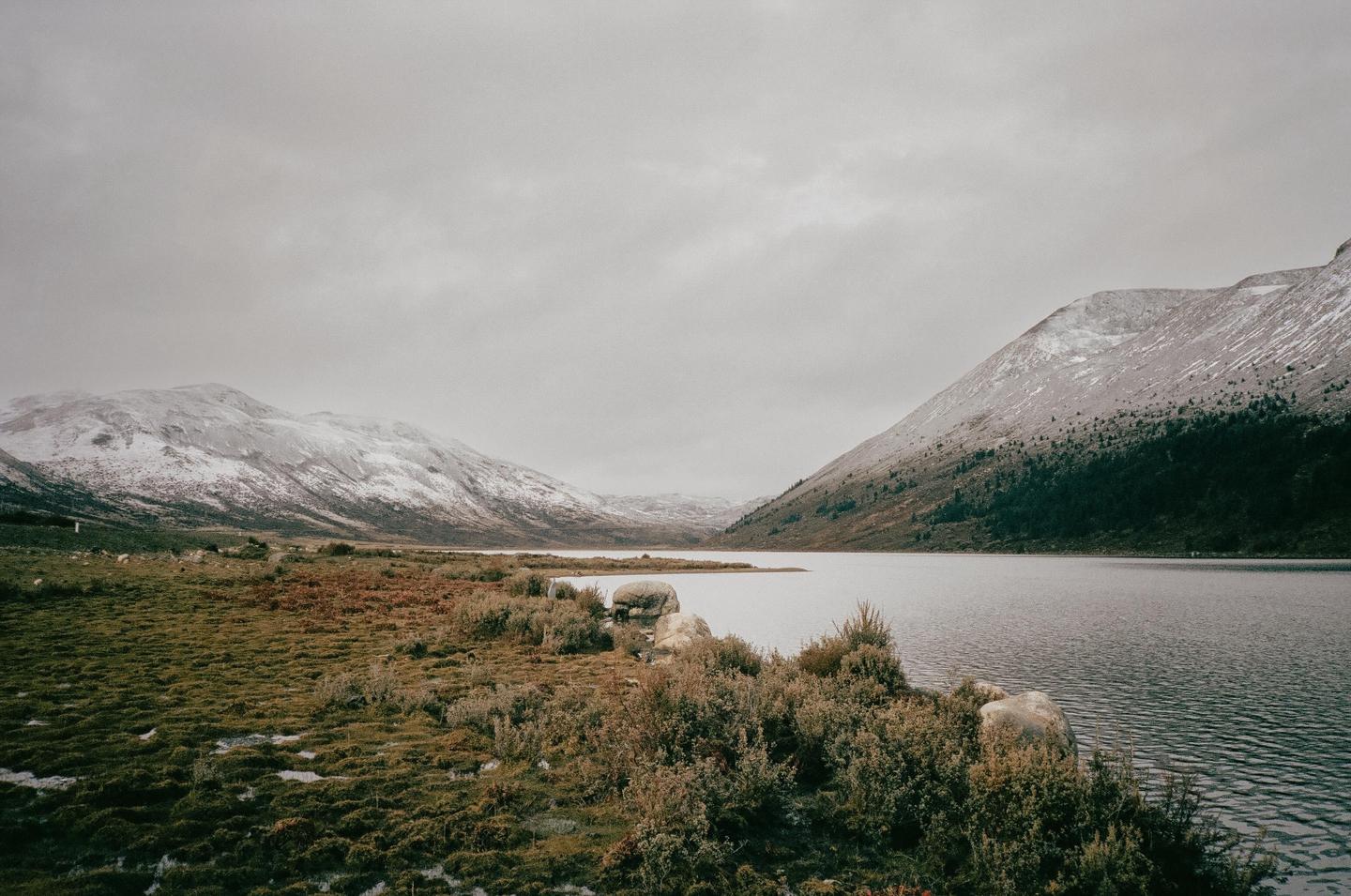 A lake is surrounded by snow-capped mountains and green and brown foliage under a cloudy sky.