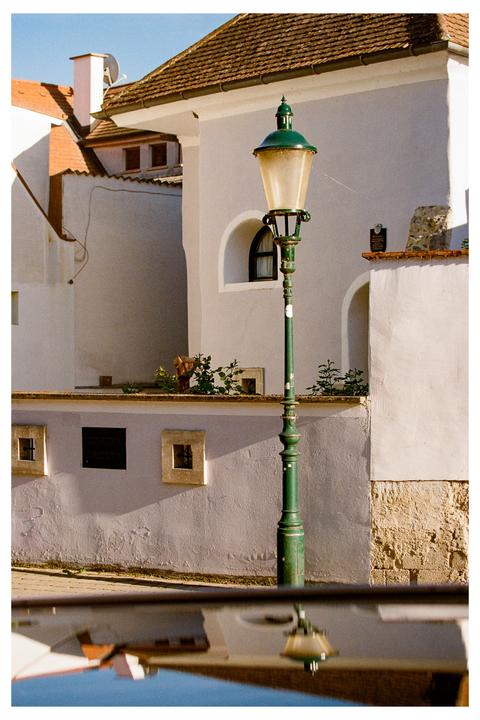 A green street lamp stands in front of a white building with a tiled roof, with its reflection visible in a surface below.