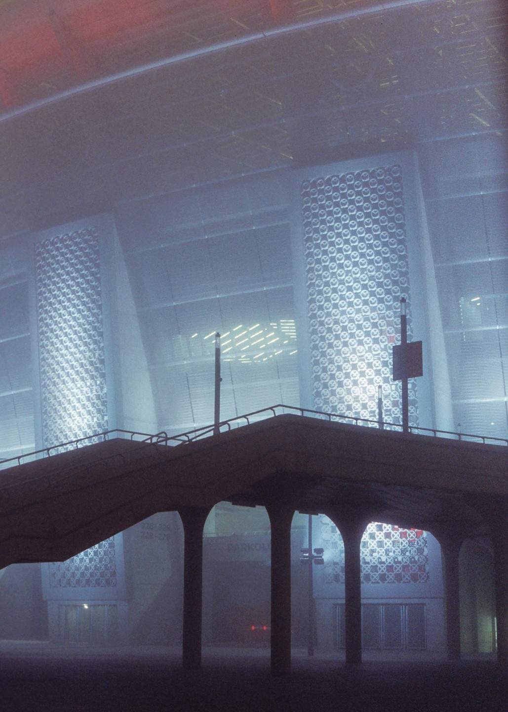 A concrete overpass with arches stands in front of a large building with patterned windows on a foggy night.
