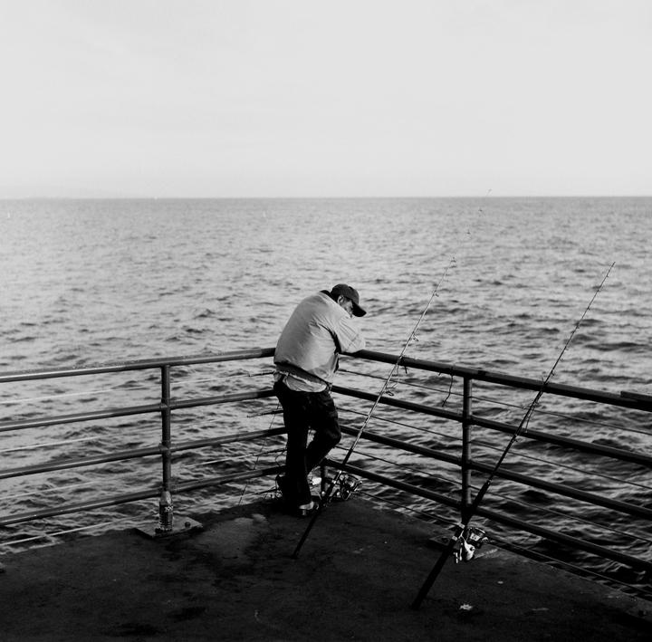 A man leans on a railing next to two fishing rods overlooking the ocean.
