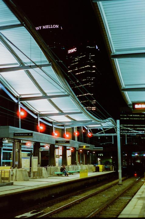 A person sits on a bench at a train station platform at night with a city building in the background.