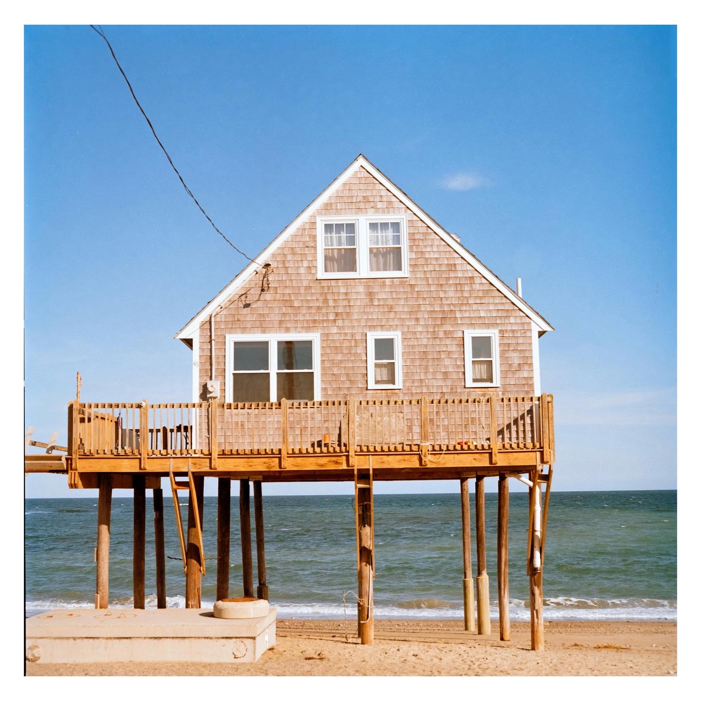 A house on stilts stands on a sandy beach with the ocean in the background.