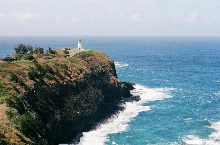 A white lighthouse with a red roof stands on a green and brown cliff overlooking a blue ocean with white waves.