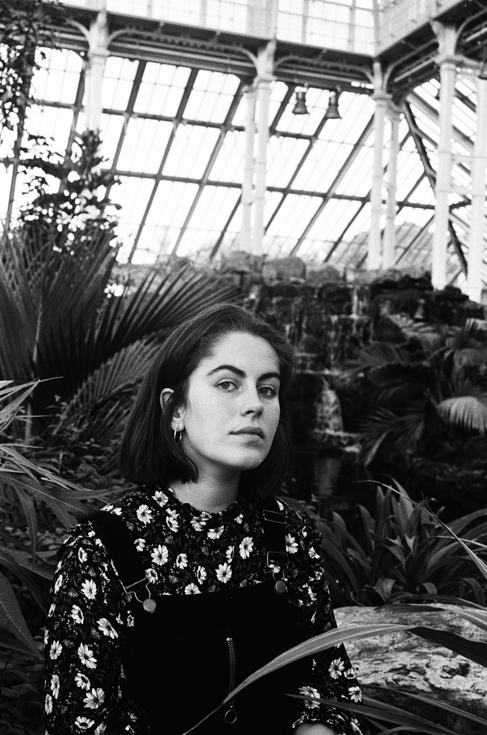 A woman in a floral shirt and overalls stands among plants in a greenhouse.