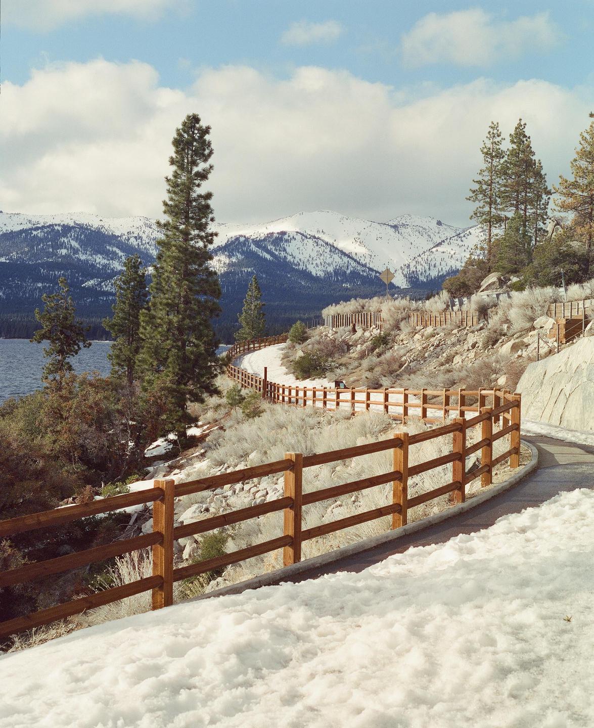 A winding path with a wooden fence runs alongside a lake, with snow-capped mountains and pine trees in the background.