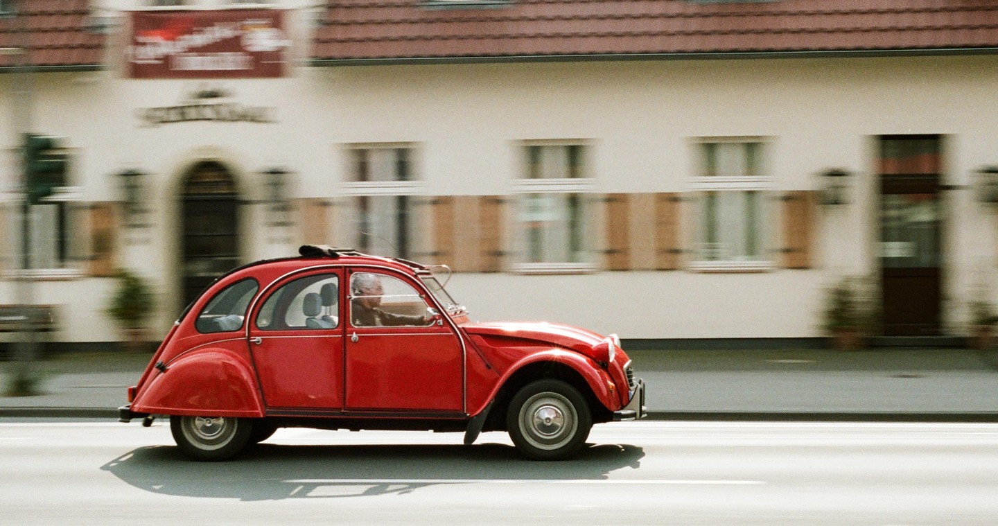 A red car with a man driving is in motion on a street with a building in the background.