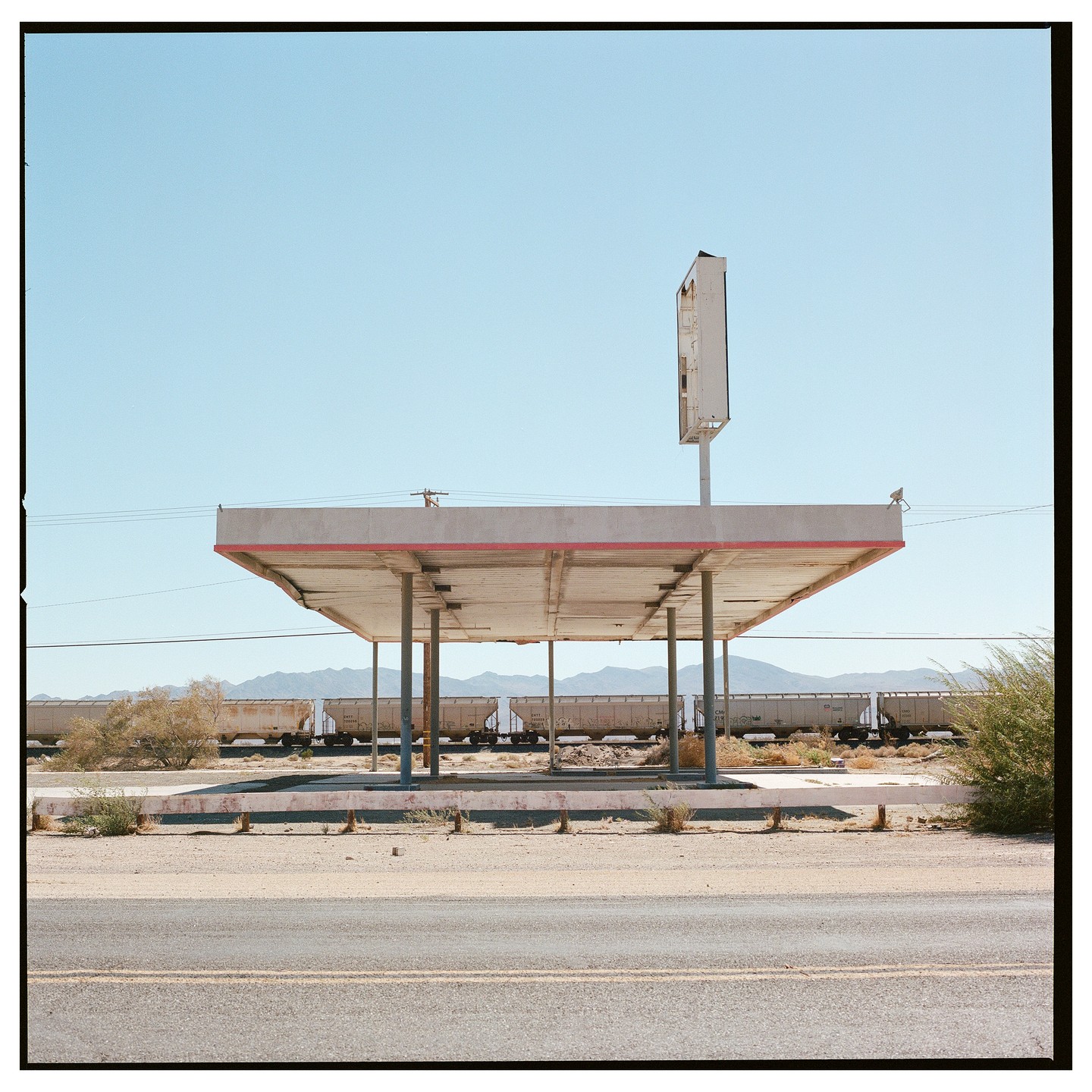 An abandoned gas station canopy stands in a desert landscape with a train passing in the background.