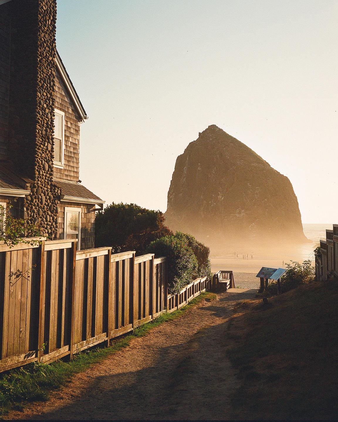 A dirt path leads to a beach with a large rock formation in the distance, next to a house with a stone chimney and a wooden fence.