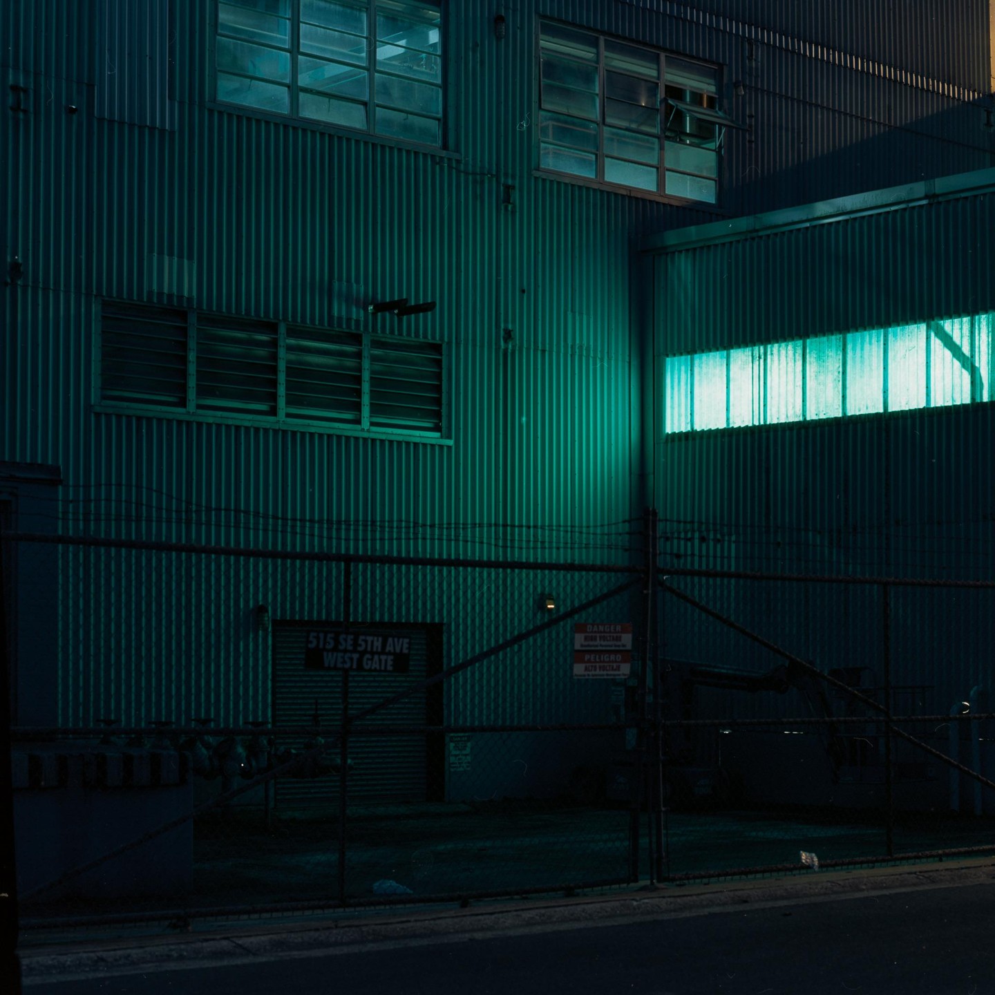 A corrugated metal building with glowing windows is seen behind a chain-link fence at night.
