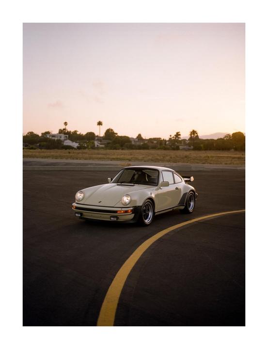 A cream-colored vintage car with its headlights on is parked on an asphalt road with a yellow line, with palm trees and buildings in the background under a sunset sky.
