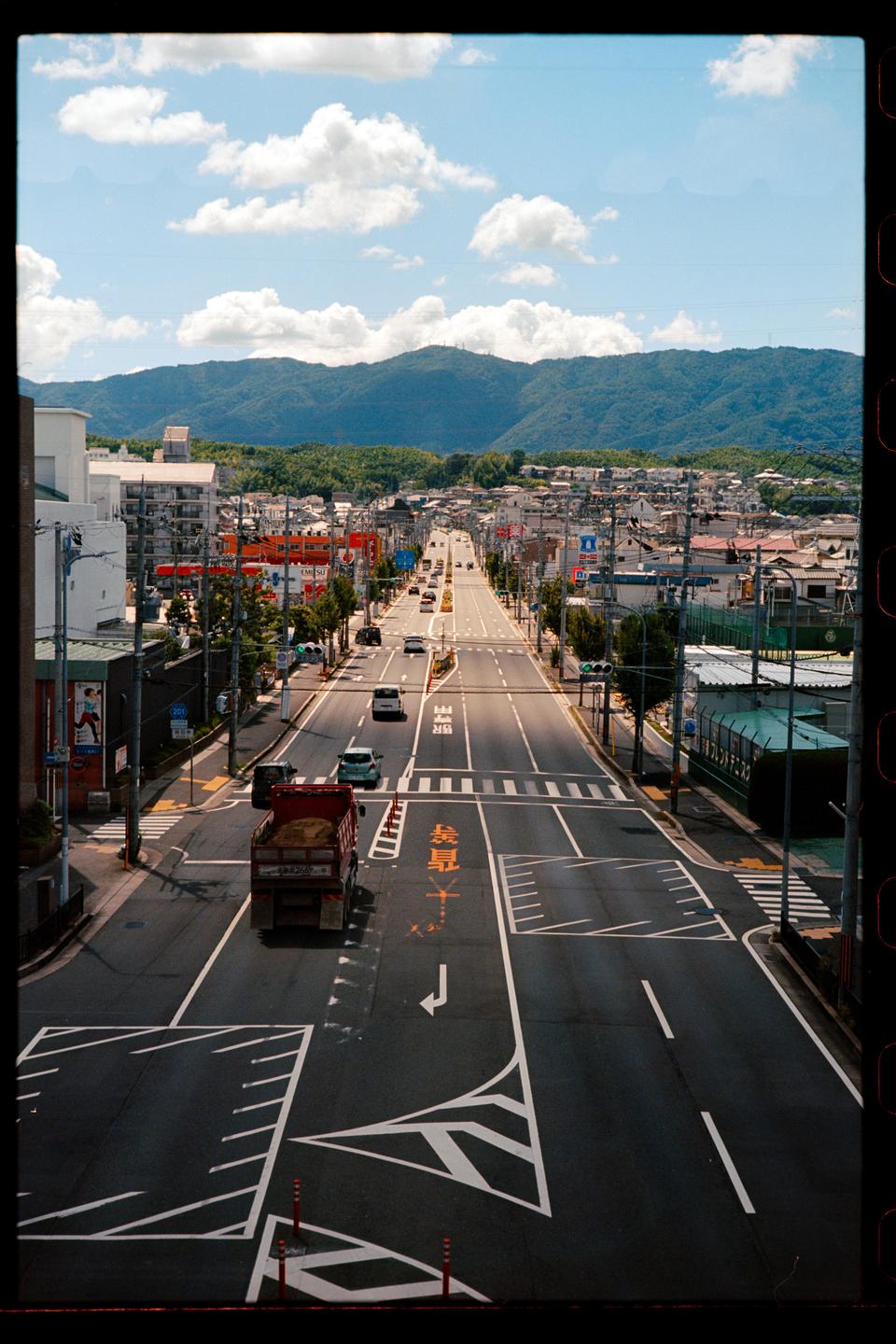 A wide street with cars and buildings on either side leads towards distant mountains under a blue sky with clouds.
