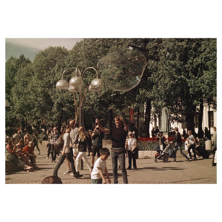 A man makes a large bubble in a park with many people and trees in the background.