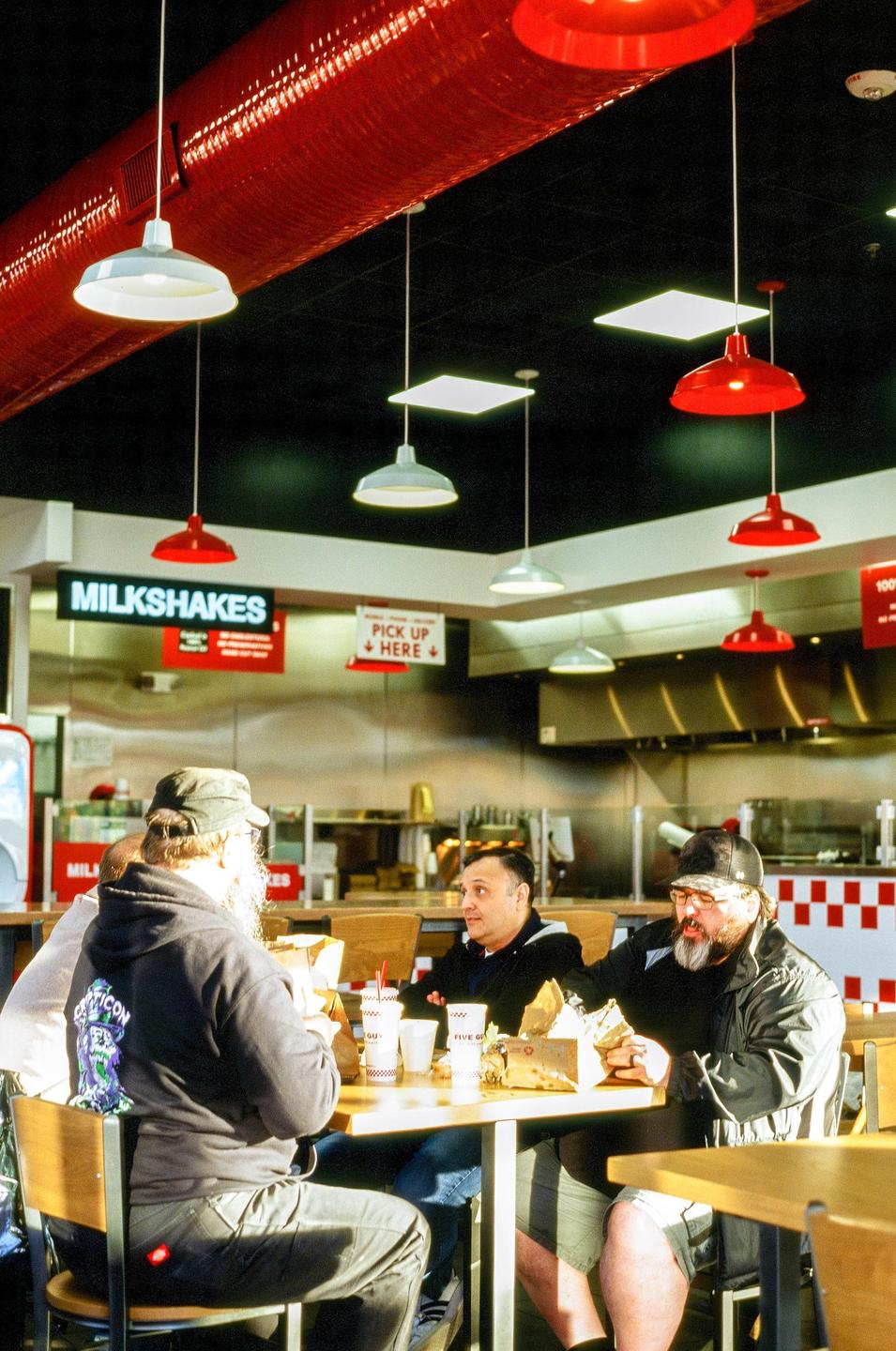 Four men are seated at a table in a restaurant, eating burgers and fries under red and white hanging lamps.