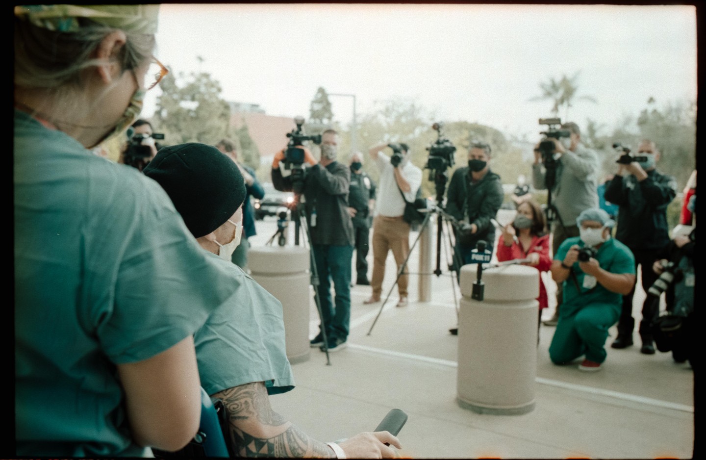 A man in a wheelchair with a tattooed arm is surrounded by medical staff and news reporters with cameras.