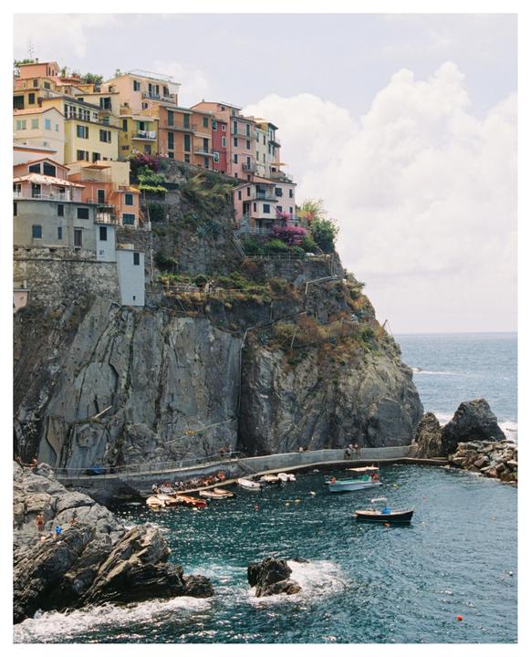 Colorful buildings are perched on a cliff overlooking a small harbor with boats and people.
